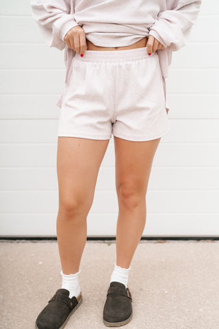 A person wearing light pink textured shorts, a matching light pink sweatshirt, white socks, and dark brown slip-on shoes, standing in front of a white garage door.