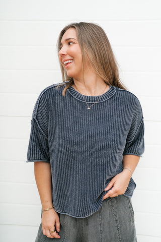 A woman wearing a dark navy blue textured knit short-sleeve top and gray jeans, standing against a white shiplap wall.