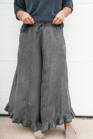 A person wearing wide-leg charcoal gray ruffled pants with a drawstring waist, paired with a dark blue ribbed top and beige shoes, standing against a white paneled wall.