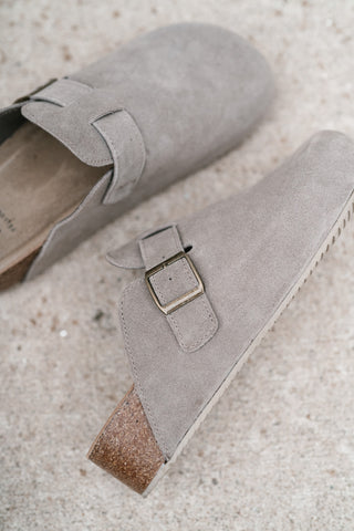 A pair of taupe suede clogs with a single strap and a gold-tone buckle, resting on a textured concrete surface.