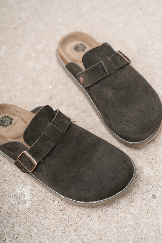 A pair of dark brown suede clogs with adjustable straps and metal buckles, displayed on a textured beige stone surface.