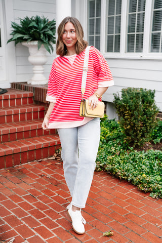 A woman wearing the pink and red horizontal striped oversized short sleeve top paired with light-wash denim jeans and white sneakers on a brick path.