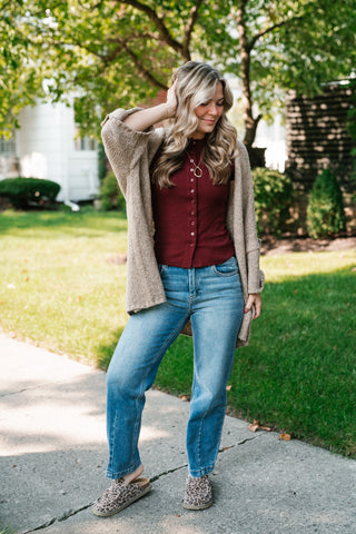 A woman stands on a concrete path in a grassy backyard, wearing a beige knit cardigan over a burgundy top, blue jeans, and leopard print slip-on shoes.