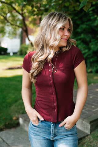 A woman with blonde hair wearing a short-sleeve maroon button-up shirt and blue jeans, standing outdoors in a garden setting.