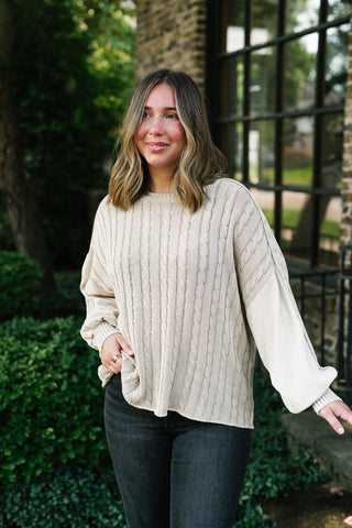 A woman wearing a cream cable-knit sweater with contrasting solid knit sleeves, paired with dark gray jeans, standing outdoors in front of a brick building and greenery.