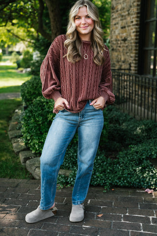 A woman stands outdoors wearing a dusty rose cable-knit sweater, blue jeans, and gray slip-on shoes, with greenery and a stone building in the background.