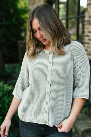 A woman wearing a light sage green ribbed knit cardigan with a button-down front, photographed outdoors in front of a rustic brick building.