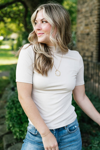 A woman wearing a fitted short-sleeve cream mock neck top and blue jeans, standing outdoors in front of a stone wall and greenery.