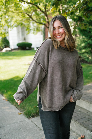 A woman wearing an oversized, faded brown knit sweater and dark jeans, standing outdoors on a paved path with a grassy lawn and trees in the background.