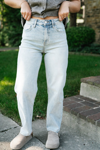 A person wearing light-wash, high-rise straight-leg jeans, a gray cropped cardigan, and beige slip-on shoes, standing outdoors on a stone path with green grass and brick steps in the background.