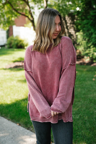 A woman wearing a dusty rose pink oversized knit sweater with decorative frayed trim details on the sleeves, standing outdoors on a concrete path with a green grassy lawn and trees in the background.