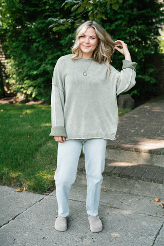 A woman stands outdoors on a paved walkway wearing an oversized sage green long-sleeve top, light blue jeans, and taupe slip-on shoes, with green trees and grass in the background.