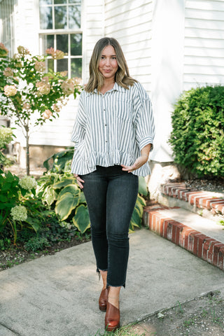 A woman stands on a concrete walkway in front of a white house, wearing a light blue and white vertically striped long-sleeve blouse with a ruffled hem, dark gray skinny jeans, and brown leather ankle boots.