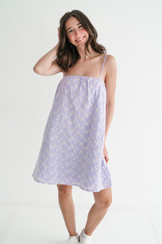 Smiling woman wearing a lavender floral eyelet mini dress with spaghetti straps against a white studio background.