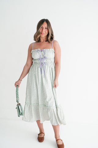 Woman wearing a sage green and purple floral midi dress with a lace-up bodice and tiered skirt, holding a green braided handbag.