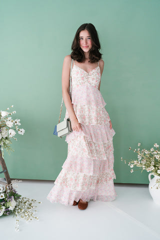 Woman in a pink and white tiered floral maxi dress with spaghetti straps, posing against a soft green background with spring flowers.