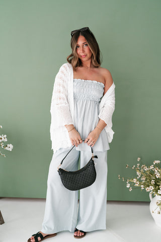 Woman wearing a light blue smocked tube top and matching wide-leg pants with a white knit cardigan and black handbag.