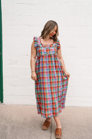 A woman modeling the Oksana Rainbow Plaid Midi Dress featuring vibrant multi-colored checks, ruffle sleeves, and a square neckline against a white brick background.