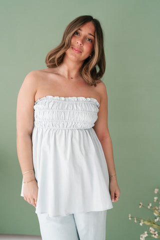 Woman modeling a light blue smocked strapless peplum top with ruffle detailing against a sage green background.