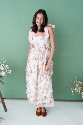 Woman smiling in a white floral smocked maxi dress featuring pink bow shoulder straps and tiered ruffles against a sage green background.