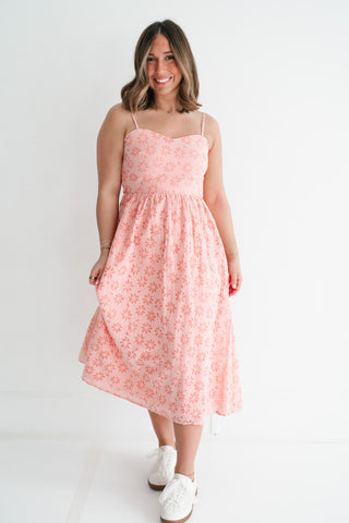 Woman wearing a coral and white gingham floral midi dress with spaghetti straps and white sneakers against a white background.