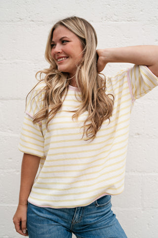 Blonde woman smiling in a yellow and white striped Daydreamer oversized tee with pink contrast trim and blue jeans against a white brick wall.