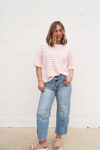 Model wearing the pink and white Daydreamer Oversized Contrast Stripe Tee with light-wash denim jeans and neutral sandals against a white brick wall.