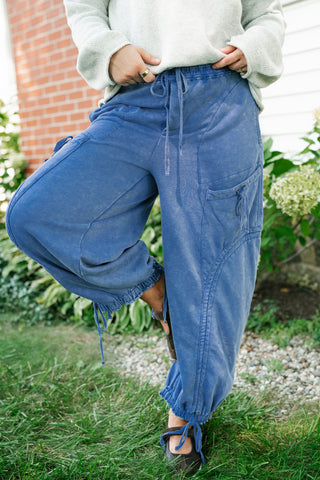 A person outdoors wearing loose blue drawstring pants with elastic cuffs, paired with a light gray sweatshirt and dark lace-up shoes, standing on grass near a brick wall and garden plants.