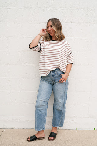 Woman wearing the Daydreamer mocha and white striped oversized tee with dark contrast trim and light-wash denim jeans against a white brick wall.