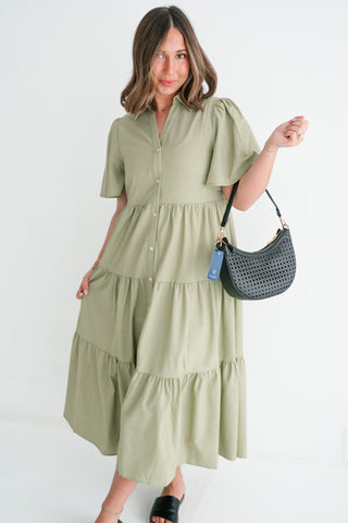 Woman wearing a sage green tiered midi dress with flutter sleeves and a button-down front, paired with a black woven shoulder bag against a white background.