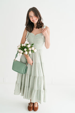 Woman wearing a sage green floral tiered midi dress with a smocked bodice and tie-up straps, holding white roses and a matching green handbag.