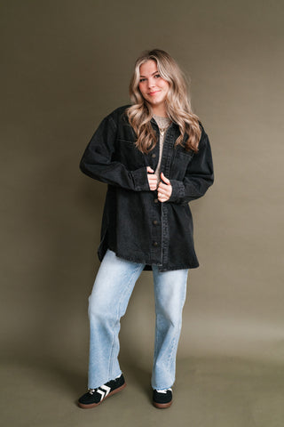 Woman wearing a black washed denim button-front shacket layered over a neutral top with light-wash straight-leg jeans and black sneakers.