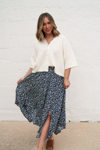 A woman wearing the Sunny May Ivory Knit Button-Up cardigan with wide 3/4 sleeves, featuring a boxy fit and rolled collar, paired with a navy floral midi skirt.