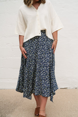 A woman wearing the Serenity Navy Floral Midi Skirt, featuring a ditsy floral print, handkerchief hem, and paired with a white button-up cardigan.