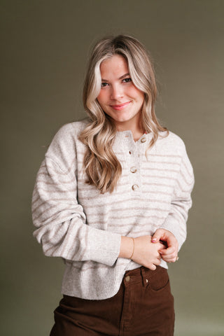 Woman wearing a soft oatmeal and beige striped knit henley sweater with a button neckline, long sleeves, and ribbed trim, styled with high-waisted chocolate brown pants.