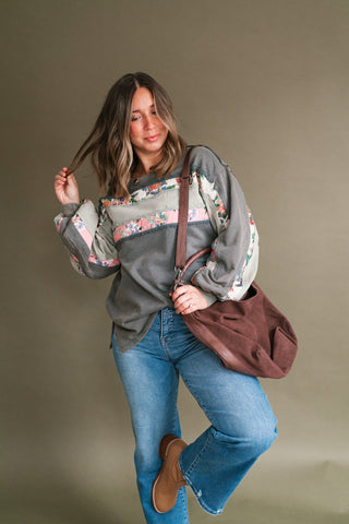 A woman posing in a gray long-sleeve sweatshirt with floral patterned sleeves and a brown suede shoulder bag, paired with blue jeans and brown boots, against a solid taupe background.