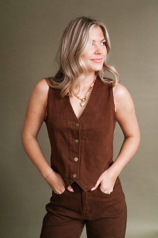 Woman wearing the West End Button Vest in rich chocolate brown with a V-neckline and gold-tone buttons, styled with matching brown pants and a beaded heart necklace.