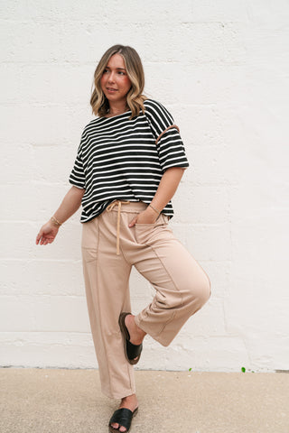 A woman wearing the Daydreamer oversized black and white striped tee with a mocha contrast crew neckline tee, paired with tan joggers and black sandals against a white brick wall.