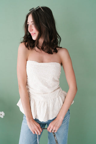 Woman wearing a cream eyelet strapless peplum top and blue jeans against a green background.