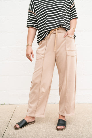 A woman wearing mocha-colored utility barrel leg pants with an elastic drawstring waistband, exposed seam detailing, and large front pockets, styled with a striped tee and black slides.