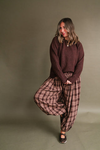 Woman wearing a deep chocolate brown long sleeve knit sweater styled with brown and mauve plaid balloon pants, layered gold necklaces, and brown sneakers.
