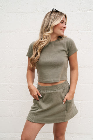 A model wearing the sage green "Weekend Feeling" ribbed knit set featuring a short-sleeve crop top and matching skort with deep front pockets.