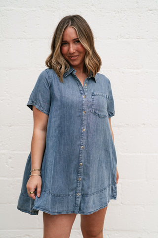 A woman wearing the Core Classic Tencel Chambray Mini Dress, featuring a light-wash denim look, button-front design, and a relaxed fit against a white brick background.