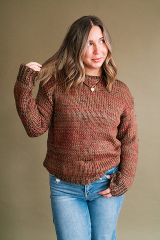 Woman wearing the Golden Ridge Long Sleeve Knit Sweater featuring a chunky multicolor rust, olive, and brown knit, styled with light wash jeans and layered gold necklaces.