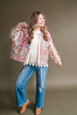 A woman modeling a colorful bohemian floral print bomber jacket over a white lace-trim top, blue jeans, and tan boots, set against a solid taupe studio background.