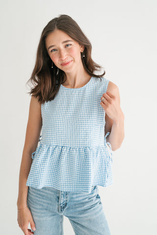 Woman wearing a sky blue and white gingham peplum top with side-tie details and denim jeans against a white background.