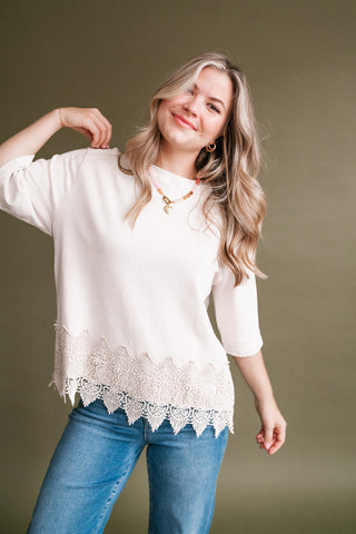 Woman wearing an ivory mineral-wash short sleeve top with a detailed floral lace hem, styled with blue jeans.