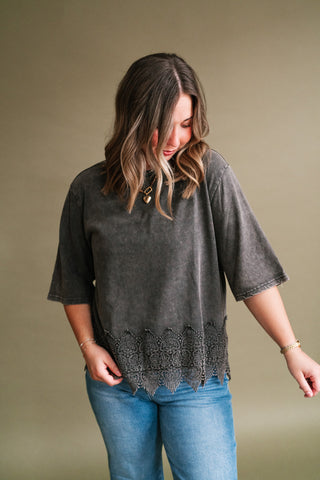 Woman wearing a charcoal mineral-wash short sleeve top with a detailed floral lace hem and blue jeans.