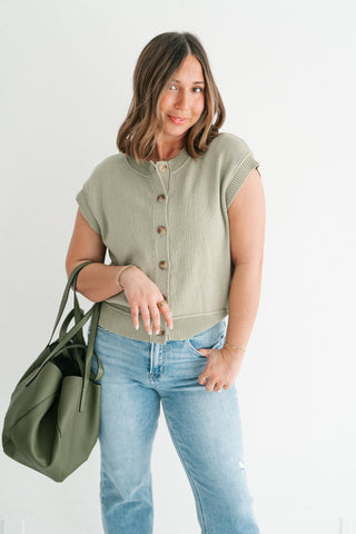 Woman wearing a sage green cap-sleeve button-down knit vest paired with light wash jeans and a large olive green leather tote bag.
