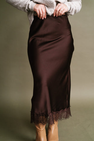 Woman wearing a chocolate brown satin midi skirt with delicate lace trim along the hem, styled with a soft neutral cardigan and tan boots.